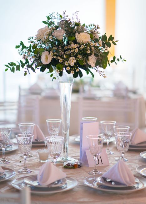 A tall, elegant glass vase holding a lush bouquet of fresh white roses, small white daisies, and sprigs of greenery with dark green leaves, arranged in a rounded, loosely gathered style. The floral ar