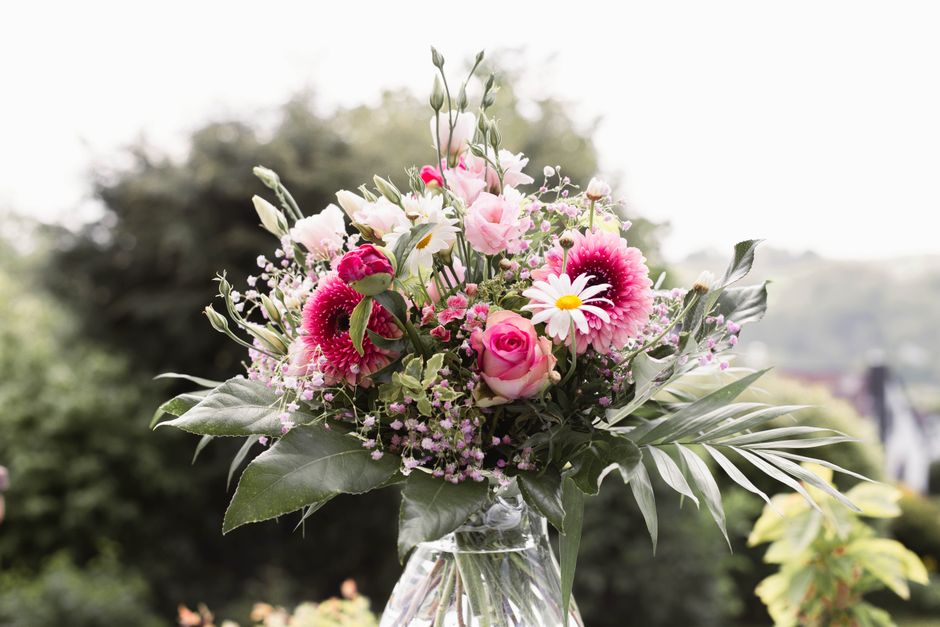 A vibrant floral bouquet featuring pink roses, white daisies with yellow centers, pink and white gerbera daisies, and small pink filler flowers, arranged with lush green foliage including large, elong