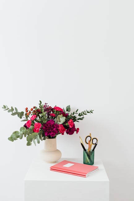 A floral arrangement featuring a mix of vibrant pink and deep magenta carnations and roses, complemented by sprigs of greyish-green eucalyptus leaves, all arranged in a smooth, white ceramic vase plac