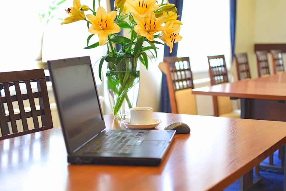 A clear glass vase filled with fresh yellow lilies and green foliage sits on a polished wooden conference table, with the flowers arranged in a loose, natural bouquet. The lilies' petals are vibrant a