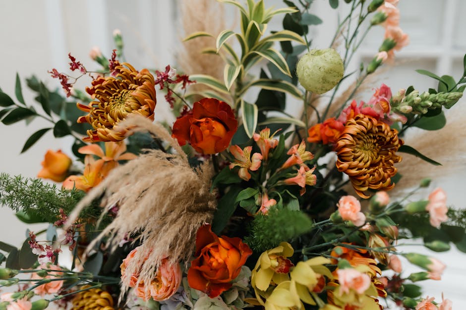 An arrangement of fresh flowers featuring orange roses, peach and pink snapdragons, and large amber chrysanthemums, complemented by green foliage including variegated leaves and delicate sprigs. The b