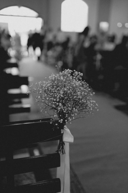 A delicate bouquet of baby's breath flowers with tiny white blooms and green stems, tied with a satin ribbon, is attached to the back of a dark wooden chair in an indoor setting. The background shows 